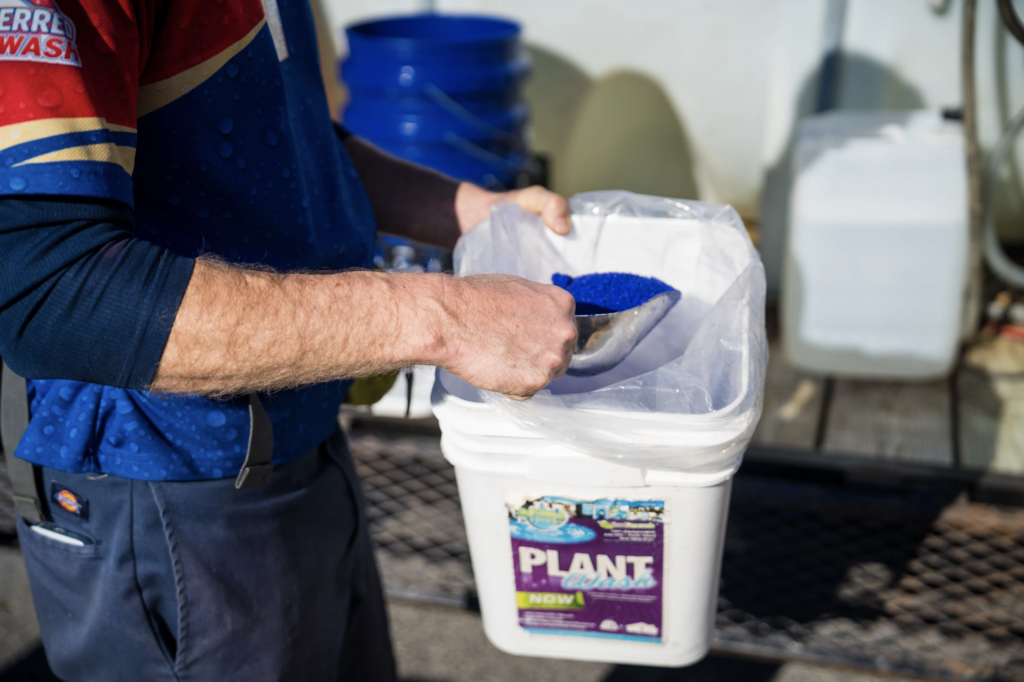 A person is scooping blue granular substance from a white plastic container labeled 